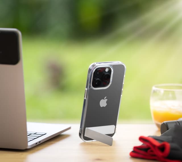 Aero clear case holding an iPhone upright on a wooden table with its kickstand bar swung out, laptop and juice glass nearby in a sunlit garden