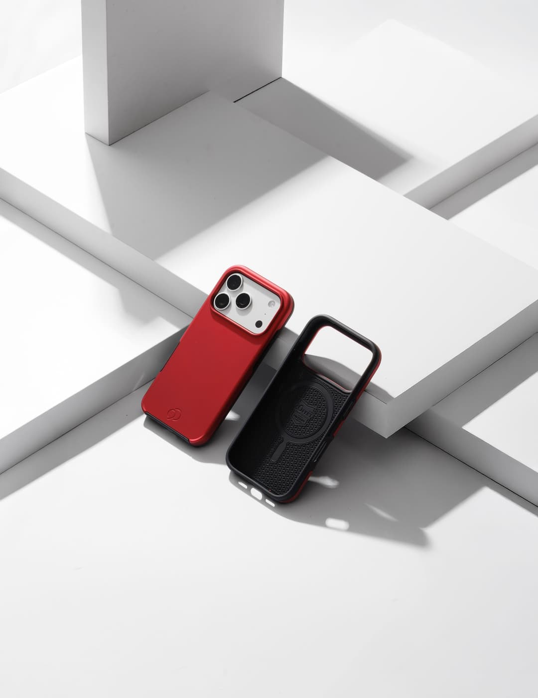 Crimson Cirrus 3 case on a white iPhone, stood beside a black Cirrus 3 shell that shows its ridged interior and magnet ring, on white studio blocks