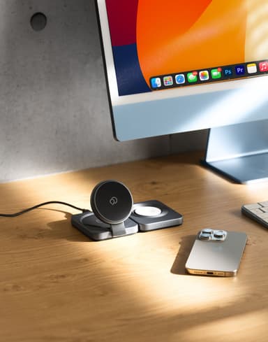 The Qi2 charging station on a wood desk in front of a monitor, its magnetic pad raised and the round watch charger beside it