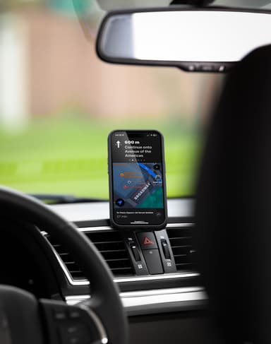 A phone standing in portrait on the black Nimbus9 dash mount running turn-by-turn navigation, seen from the driver seat with green trees blurred through the windshield