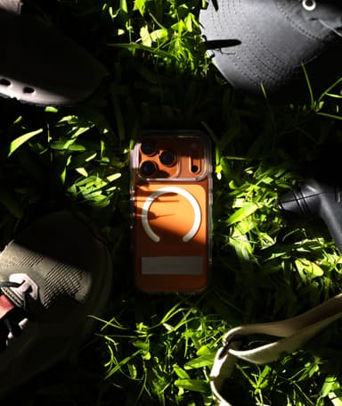 Clear Aero case on a cosmic-orange iPhone 17 Pro lying in sunlit grass, MagSafe ring and closed kickstand bar showing through the shell