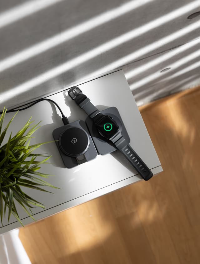 Overhead view of the charging station lying open on a white surface with its magnetic pad folded flush, an Apple Watch charging on the panel beside it, venetian-blind light across the desk