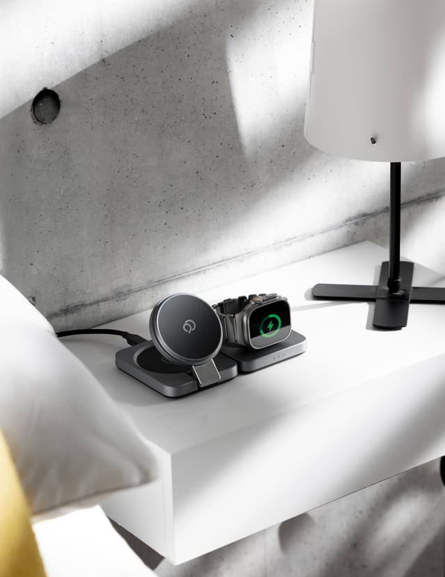 The charging station on a white nightstand, its magnetic pad raised on its hinge above the flat base pad and an Apple Watch charging on the section beside it, a slim lamp and a concrete wall behind