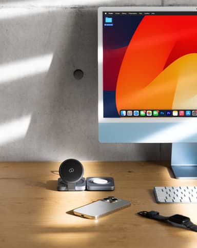 The charging station on a wood desk in front of a monitor, a phone and a watch lying beside it, sunlight striping the concrete wall behind