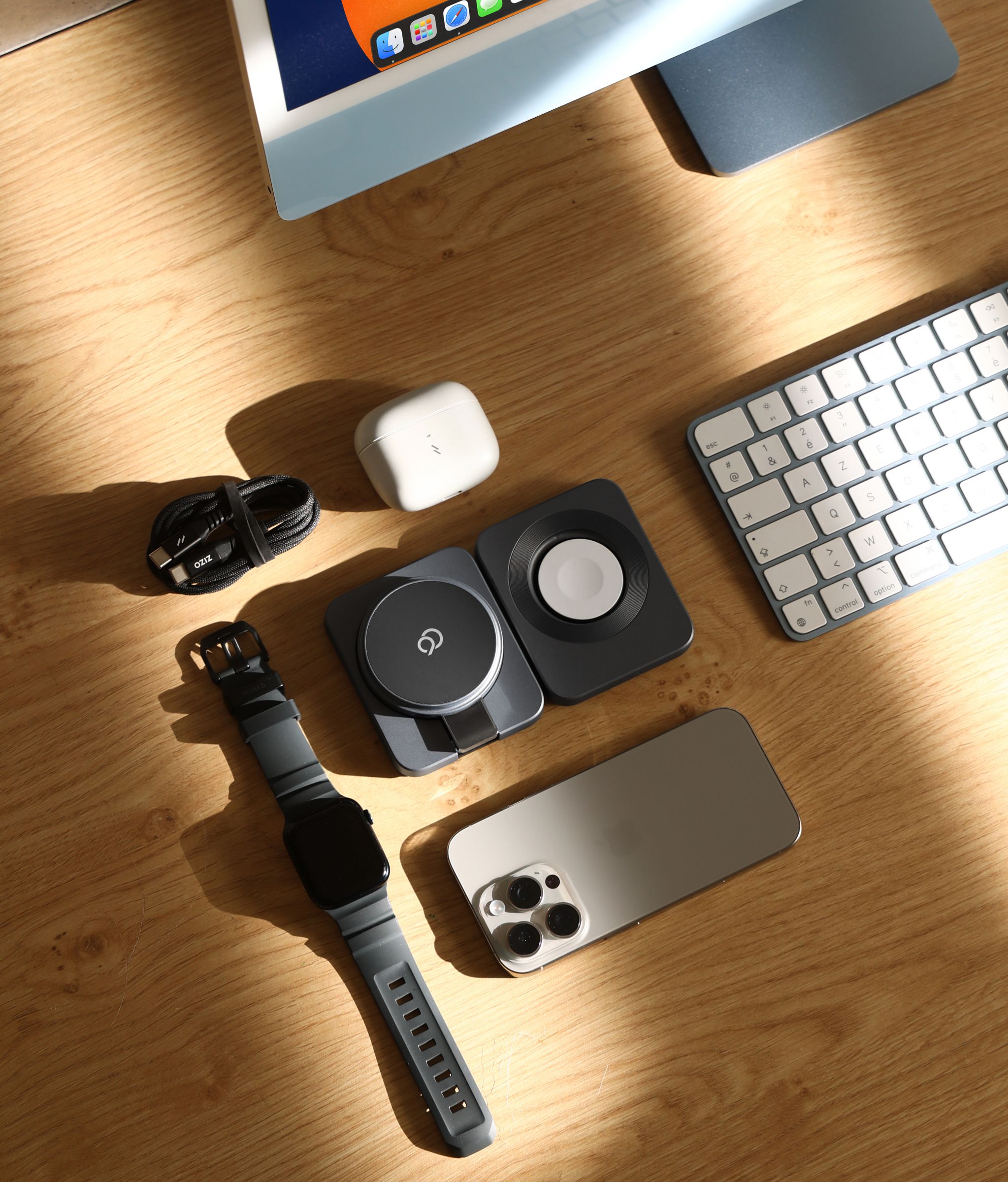 Overhead view of the charging station open flat on a wood desk, with a phone, an Apple Watch, an earbuds case, a coiled cable and a keyboard laid out around it