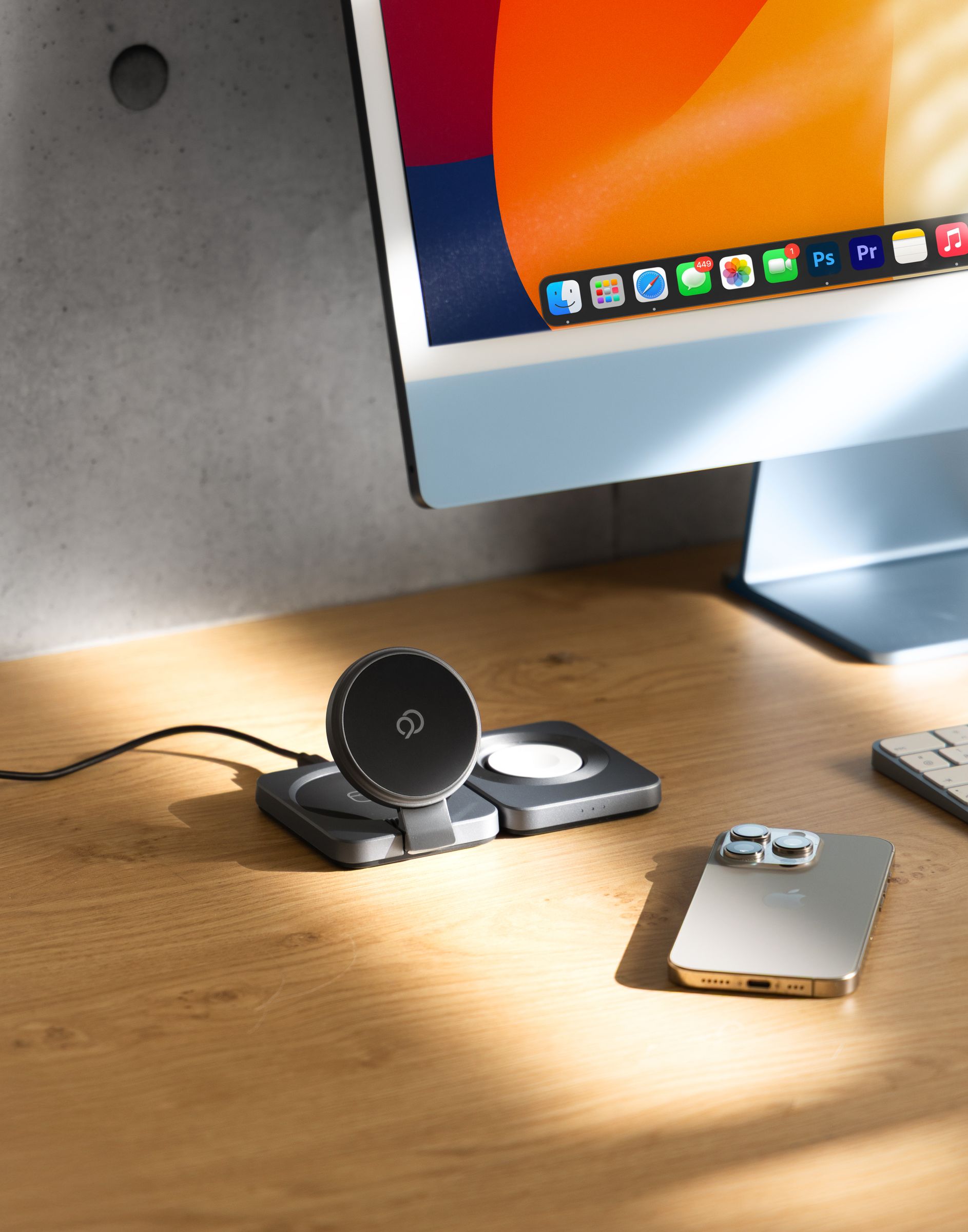 The charging station on a wood desk in front of a monitor, its magnetic pad raised and the round watch charger beside it, a phone lying face down to the right and the cable running away to the left