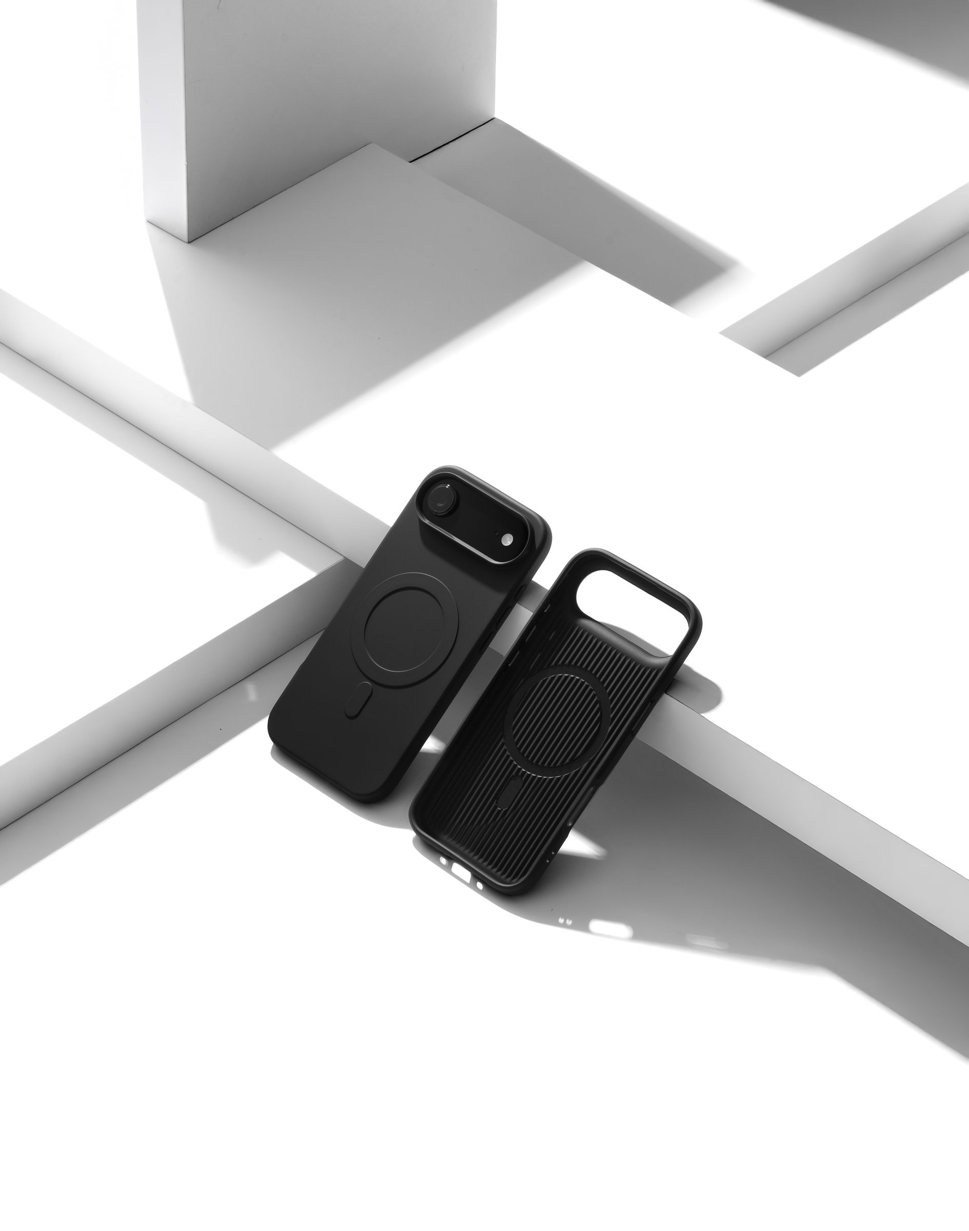 Two Nimbus9 Alto 2 cases in Black on a white geometric studio set — one fitted on a phone, one propped empty with its MagSafe ring showing