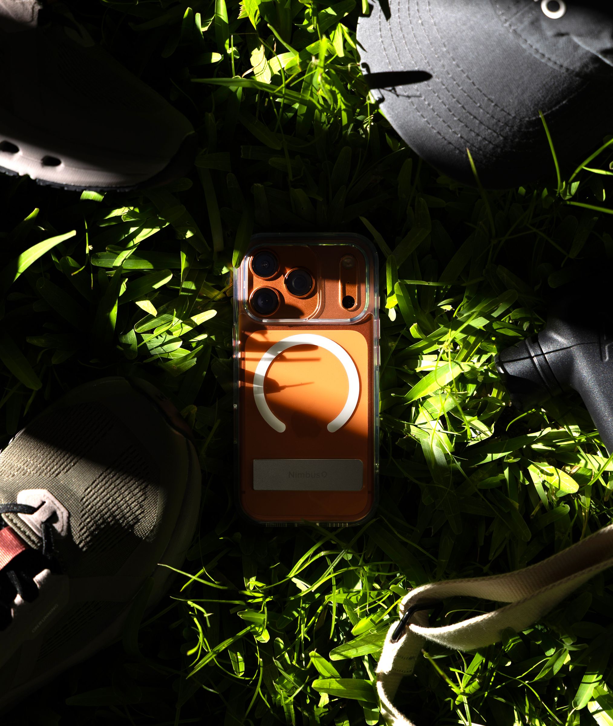 Clear Aero case on a cosmic-orange iPhone 17 Pro lying in sunlit grass, MagSafe ring and closed kickstand bar showing through the shell