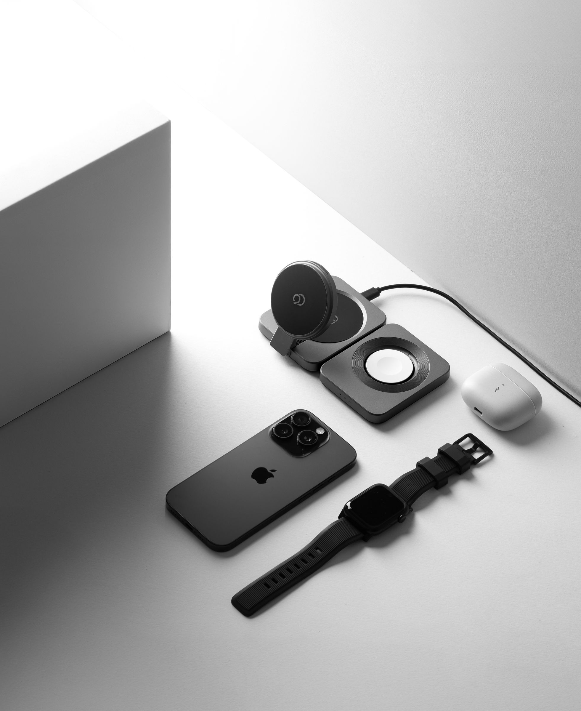 The charging station open on white, its magnetic pad raised and the round watch charger in the section beside it, with a phone, a watch and an earbuds case laid out on the surface, none of them docked