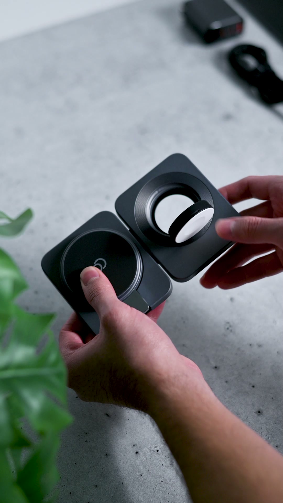 Two hands holding the brushed gray charging station open, a flat charging pad on one panel and the round watch charger sitting in its cradle on the other, over a concrete desk